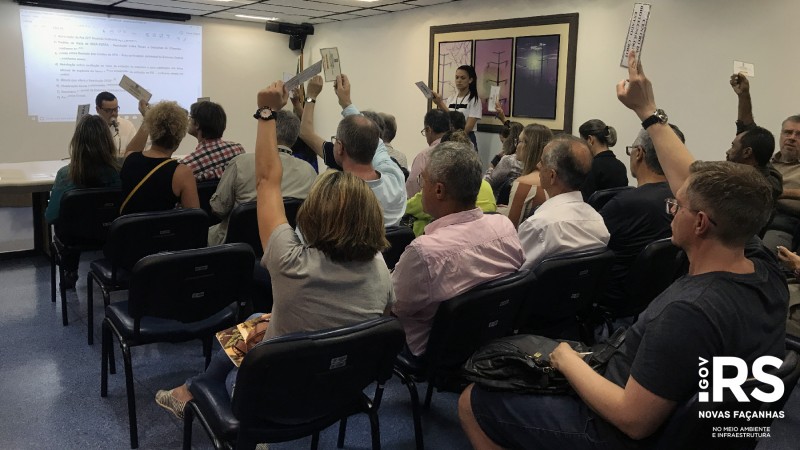 A imagem retrata várias pessoas sentadas em uma sala de reuniões. Algumas delas estão com o braço levantado e seguram um papel para votação.