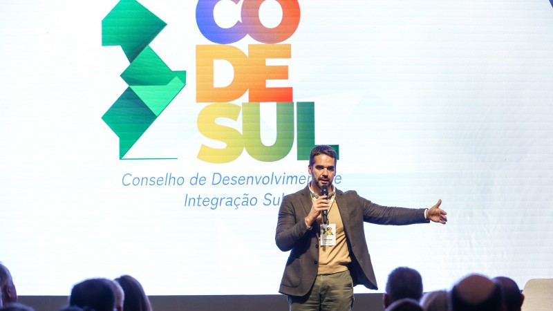 Governador no palco falando para a plateia. Ao fundo, painel de led com a logomarca do Codesul nas cores verde, laranja e vermelho.