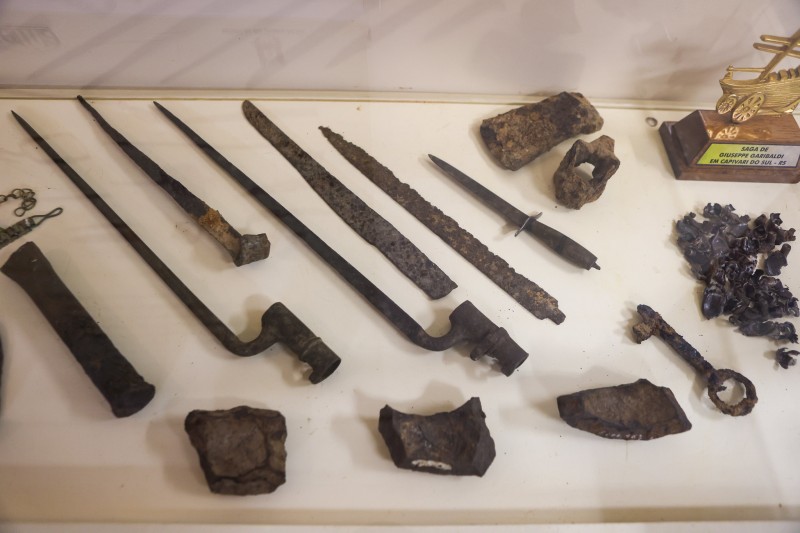 em torno de 10 artefatos enferrujados e deteriorados pelo tempo sobre uma mesa branca. Entre eles, ponta de lança, chave e fragmentos de metal nao-identificados