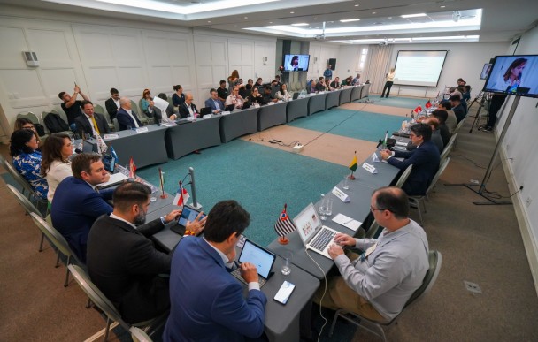 Plano geral dos secretários sentados no entorno de uma mesa em formato de ferradura.