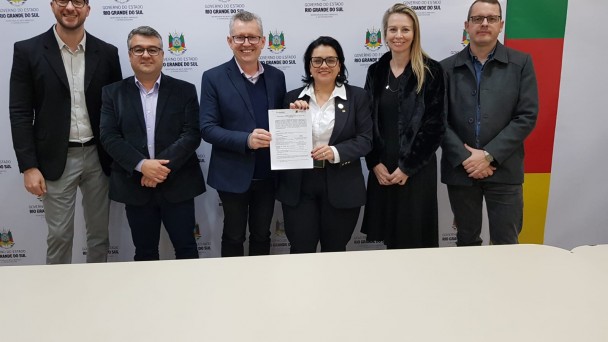 Secretários da Sema , presidente da Fapergs e a equipe da Asclima em pé.Ao fundo o painel branco com brasões do Estado. No centro, a secretária da Sema e o presidente da Fapergs seguram o Edital de monitoramento.