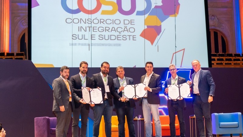 Governadores em pé no palco segurando o documento do tratado. Ao fundo a logomarca Cosud nas cores azul, laranja, amarelo e roxo.