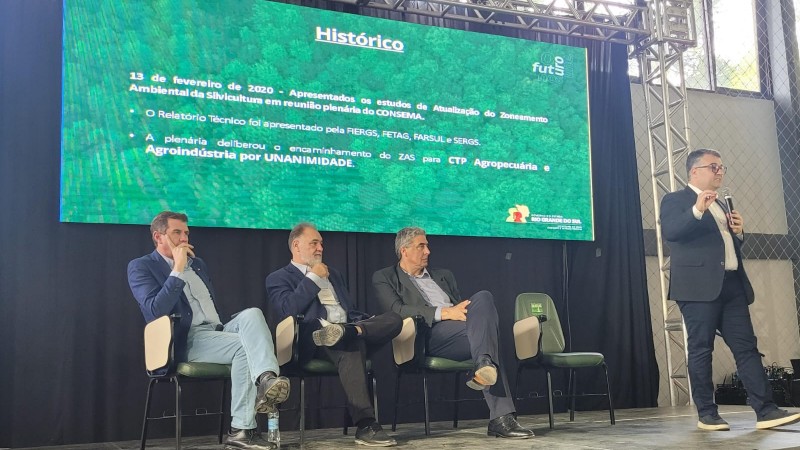 Três homens sentados em linha e um em pé apresentando o painel que está com imagens dispostas ao fundo do palco em um telão.