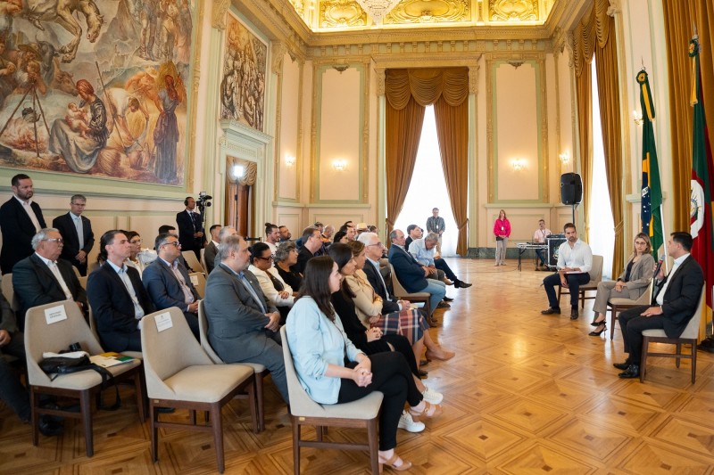 Imagem aberta do Salão Alberto Pasqualini, no Palácio Piratini. De perfil podem ser vistos, à direita da imagem e sentados, deputados que participaram da reunião. Mais à direita está Leite, sentado, de frente para imagem, enquanto acompanha a fala de outra pessoa que está de perfil. Há ainda uma terceira pessoa sentada, à direita da imagem, próxima ao governador.