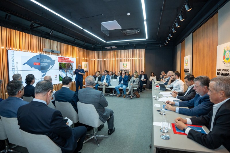 Imagem aberta da reunião realizada na Sala R1, localizada no subsolo do Palácio Piratini. À esquerda há plateia, sentada e de costas, seguida da tela e do comandante dos bombeiros, que está em pé fazendo uma apresentação, e plateia, mais distante e sentada, aparecendo de frente na imagem. Mais à esquerda, há uma enorme mesa retangular, onde estão sentados cinco homens, sendo um deles o governador Eduardo Leite.