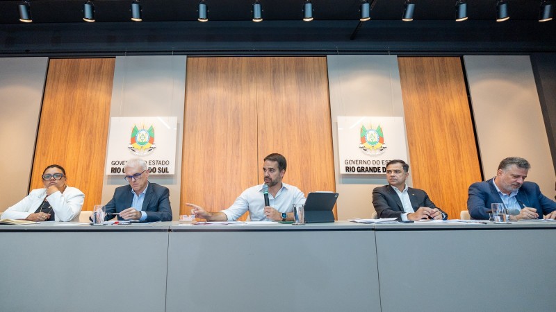 Leite sentado atrás de uma mesa, com dois homens à sua direita e dois homens à sua esquerda. O governador segura o microfone com uma das mãos e gesticula com a outra durante a sua fala.