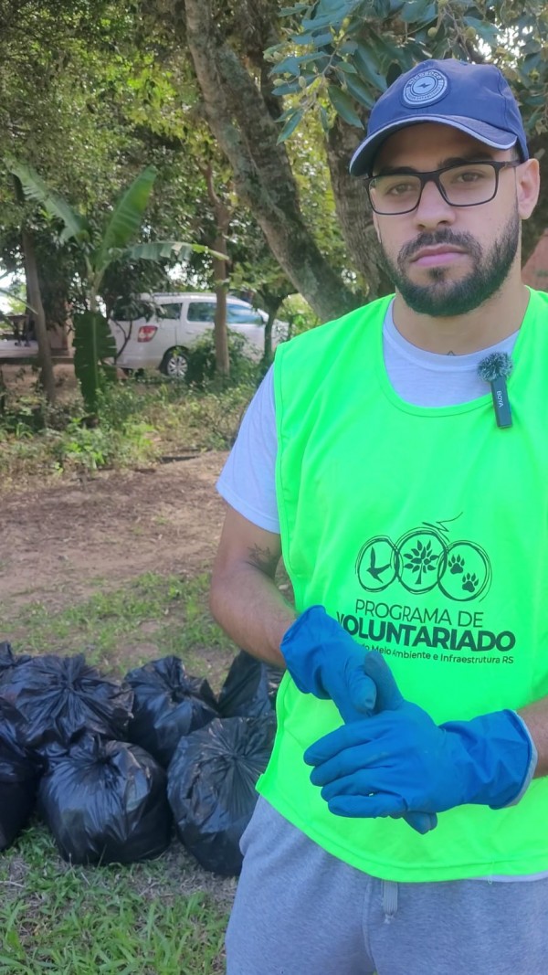 voluntário de boné e óculos de grau, luvas de borracha e colete do programa de voluntariado da sema olha para a câmera. ems egundo plano, diversos sacos contando o reculhimento dos resíduos.