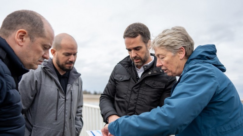 Grupo de quatro pessoas analisando um mapa ou projeto. Na esquerda, um homem de casaco preto, outro de casaco cinza, que é o secretário da Reconstrução Gaúcha, o governador Eduardo Leite, de preto, e uma senhora, mostrando o projeto, de cabelos brancos curtos e casaco azul, com capuz.