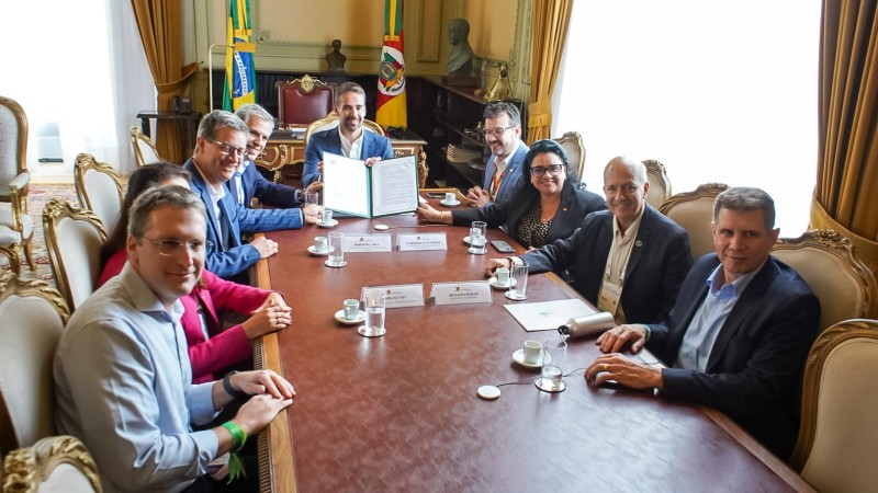 na imagem, uma mesa ao centro em primeiro plano na vertical. ao redor dela, autoridades sorriem e mostram a licença de operação entregue para a biometano sul