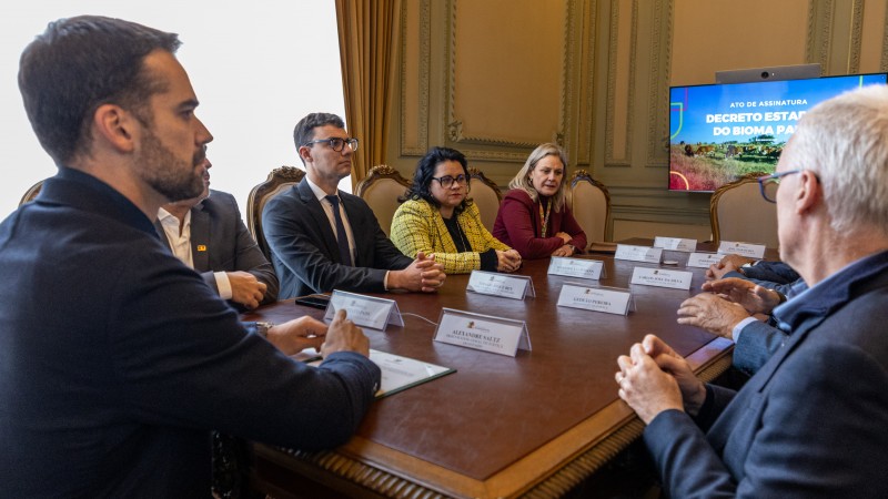 na imagem, governador aparece em primeiro plano. ao centro, uma mesa com plaquinhas brancas e, ao fundo, uma tv com o escrito: decreto estdual do bioma pampa. ao lado direito, o progurador geral alexandre saltz. do outro lado da mesa, a secretária marjorie vestida com um terno amarelo.