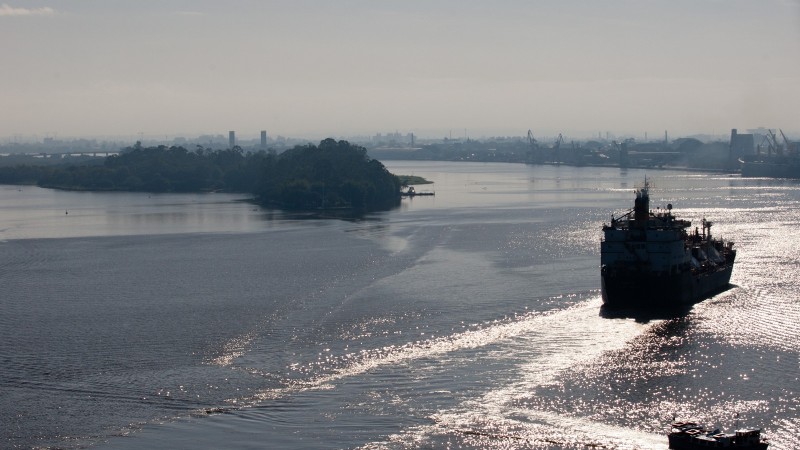 Embarcação de grande porte navega por um rio de águas calmas, com o reflexo do sol na superfície. À esquerda, uma pequena ilha coberta por vegetação. Ao fundo, estruturas industriais se destacam na linha do horizonte, parcialmente encobertas por neblina leve. O céu está claro, contribuindo para a visibilidade da paisagem fluvial.