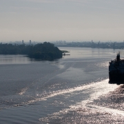 Embarcação de grande porte navega por um rio de águas calmas, com o reflexo do sol na superfície. À esquerda, uma pequena ilha coberta por vegetação. Ao fundo, estruturas industriais se destacam na linha do horizonte, parcialmente encobertas por neblina leve. O céu está claro, contribuindo para a visibilidade da paisagem fluvial.