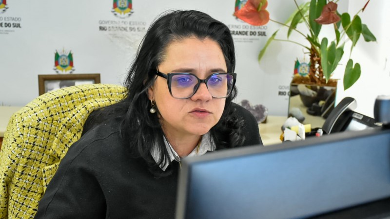 Marjorie Kaufamann, secretária da Sema, sentada em frente ao computador.Ela usa um suéter preto sobre uma camisa listrada e brincos de pérola. Ao fundo, há uma parede com o logotipo do Governo do Estado do Rio Grande do Sul, uma planta com folhas verdes e flores vermelhas, além de itens de escritório como telefone, moldura com foto e cristais decorativos sobre a mesa.