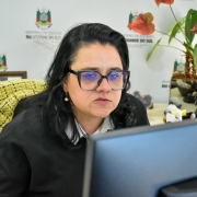 Marjorie Kaufamann, secretária da Sema, sentada em frente ao computador.Ela usa um suéter preto sobre uma camisa listrada e brincos de pérola. Ao fundo, há uma parede com o logotipo do Governo do Estado do Rio Grande do Sul, uma planta com folhas verdes e flores vermelhas, além de itens de escritório como telefone, moldura com foto e cristais decorativos sobre a mesa. 