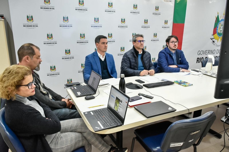 Cinco pessoas participam de uma reunião virtual sentadas ao redor de uma mesa em uma sala institucional do Governo do Estado do Rio Grande do Sul. Três homens e duas mulheres estão voltados para um monitor fora do campo de visão, aparentando atenção à transmissão. Sobre a mesa, há notebooks abertos, teclado, mouse, papéis e controles remotos. Ao fundo, há um painel com a identidade visual do Governo do RS e uma bandeira do estado parcialmente visível.