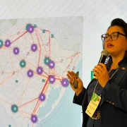 secretária marjorie, vestida de termo azul e com óculos de grau, fala ao microfone. ao fundo, um mapa do rio grande do sul.