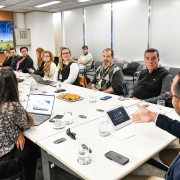 A imagem retrata uma reunião em uma sala de conferências. Diversas pessoas estão sentadas ao redor de uma mesa retangular branca, utilizando laptops, tablets ou cadernos. Sobre a mesa, há copos e garrafas de água, além de um prato com biscoitos. Ao fundo, um grande painel digital exibe a frase: "PLANO DE TRANSIÇÃO ENERGÉTICA JUSTA PARA AS REGIÕES CARBONÍFERAS DO RIO GRANDE DO SUL". O ambiente é bem iluminado por luzes fluorescentes no teto, com paredes brancas e janelas cobertas por persianas.