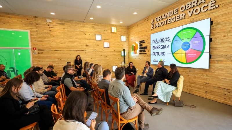 m grupo de pessoas est&aacute; reunido em uma sala com paredes de madeira clara, assistindo a um painel de debates. Quatro palestrantes est&atilde;o sentados na frente, conversando, enquanto o p&uacute;blico acompanha atento, sentado em cadeiras de madeira. Na parede, um tel&atilde;o exibe o t&iacute;tulo &ldquo;Di&aacute;logos Energia e Futuro&rdquo; ao lado de um gr&aacute;fico colorido em forma de c&iacute;rculo. No alto da parede, est&aacute; escrito em letras grandes: &ldquo;A grande obra &eacute; proteger voc&ecirc;&rdquo;.