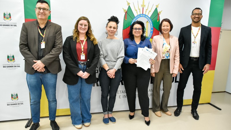 Seis pessoas posam lado a lado em frente a um painel com o bras&atilde;o do Rio Grande do Sul e as cores da bandeira do Estado. No centro, duas mulheres seguram documentos, simbolizando a assinatura de um acordo entre institui&ccedil;&otilde;es.
