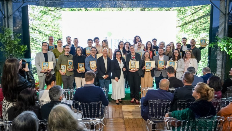 Grupo de pessoas em pé sobre um palco, segurando placas ou troféus de premiação. Ao fundo, há um painel branco com logotipo e texto parcialmente visível, indicando um evento formal de entrega de prêmios. Na frente do palco, várias pessoas estão sentadas em cadeiras transparentes, assistindo à cerimônia. O ambiente é iluminado por luz natural, com árvores visíveis ao fundo, sugerindo que o evento ocorre em um espaço aberto ou com vista para a natureza.