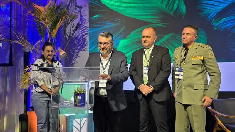 A imagem mostra quatro pessoas em p&eacute; sobre um palco, posicionadas atr&aacute;s e ao lado de um p&uacute;lpito transparente. No p&uacute;lpito h&aacute; um pequeno vaso com planta e um logotipo que indica &ldquo;5&ordm; Summit Ambiental&rdquo;. As pessoas est&atilde;o vestidas formalmente: tr&ecirc;s usam trajes sociais com crach&aacute;s pendurados no pesco&ccedil;o, e uma veste uniforme militar com ins&iacute;gnias no ombro. &Agrave; esquerda do p&uacute;lpito h&aacute; uma planta ornamental alta, e ao fundo h&aacute; um grande painel com folhas verdes e texto parcialmente vis&iacute;vel que diz &ldquo;Amor na...&rdquo;. A ilumina&ccedil;&atilde;o &eacute; intensa, com tons de azul e verde predominando no cen&aacute;rio, criando um ambiente de evento corporativo ou confer&ecirc;ncia.