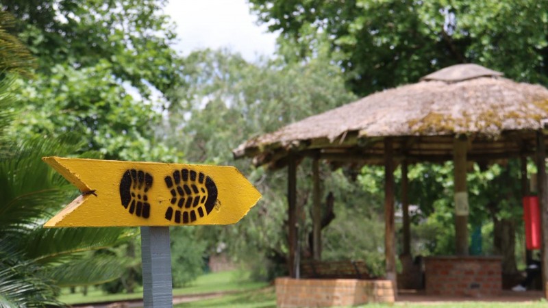 Placa amarela e com uma pegada desenhada no meio em um primeiro plano. Atr&aacute;s quiosque de madeira e vegeta&ccedil;&atilde;o verde.  