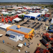 Uma fotografia a&eacute;rea colorida em plano geral e &acirc;ngulo plong&eacute;e (tomada por drone) exibe uma vis&atilde;o panor&acirc;mica e detalhada da &aacute;rea de m&aacute;quinas e implementos agr&iacute;colas da feira a c&eacute;u aberto. **O Estande Central e a Unidade M&oacute;vel:** * No centro-inferior da imagem, destaca-se uma edifica&ccedil;&atilde;o t&eacute;rrea retangular com paredes laterais laranjas e teto de zinco cinza, com um grande bal&atilde;o infl&aacute;vel esf&eacute;rico laranja fixado no topo. * Logo ao lado da estrutura, encontra-se estacionada a unidade m&oacute;vel ba&uacute; com a inscri&ccedil;&atilde;o **"CARRETAS DO SABER"** em destaque na lateral azul e branca, com toldos estendidos para recep&ccedil;&atilde;o. * Ao redor desse estande, circulam pedestres e visitantes por ruas de terra batida e cascalho claro, entre pequenas tendas de descanso com guarda-s&oacute;is brancos. **Maquin&aacute;rios e Estandes ao Redor:** * &Agrave; direita e na parte inferior, concentra-se uma expressiva exposi&ccedil;&atilde;o de pesados maquin&aacute;rios agr&iacute;colas em tons vivos: grandes pulverizadores autopropelidos e colheitadeiras vermelhas e laranjas, al&eacute;m de plantadeiras e tratores azuis e amarelos alinhados em fileiras. * &Agrave; esquerda e ao centro, estendem-se dezenas de estandes comerciais com tendas e pavilh&otilde;es modulares brancos de coberturas pontiagudas ou planas, exibindo implementos agr&iacute;colas menores, tratores, ca&ccedil;ambas e carretas vermelhas. Um dos estandes principais ao fundo traz a marca **"LS Tractor"** em um galp&atilde;o azul e branco. **O Entorno e Linha do Horizonte:** * Na parte superior da composi&ccedil;&atilde;o, al&eacute;m do limite das ruas da feira, estende-se um imenso bols&atilde;o de estacionamento com milhares de ve&iacute;culos alinhados. * Ao fundo, avistam-se &aacute;reas planas de v&aacute;rzea e espelhos d'&aacute;gua com vegeta&ccedil;&atilde;o baixa, al&eacute;m de uma alta torre/chamin&eacute; vertical &agrave; direita sob a linha do horizonte. * O c&eacute;u &eacute; limpo e azul-claro com suave bruma ao longe, iluminado plenamente pela luz do sol do meio-dia.