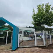 A imagem mostra a fachada mais externa da nova sede regional Sema-Fepam, em Passo Fundo. A entrada conta com um pórtico azul, sob o qual ainda há um outro telhado, esverdeado, que protege a porta de vidro. O perímetro da unidade é cercado por placas de vidro, que permitem a visualização do pátio, e uma árvore, à frente dos vidros, destaca-se. No interior, mais ao fundo, o prédio da unidade em si, com fachada branca e um telhado similar ao do acesso ao pátio cobrindo a entrada.